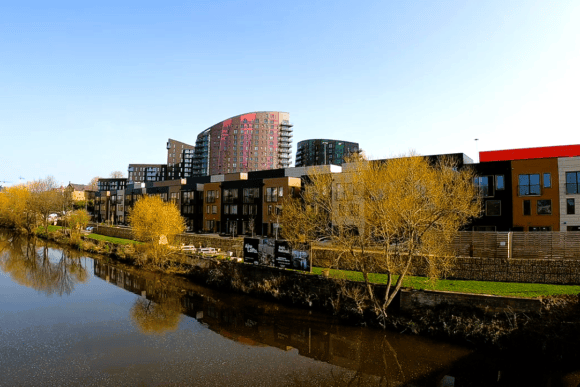 Leeds South Bank waterfront with modern apartments and riverside development reflected in the River Aire under a clear blue sky.