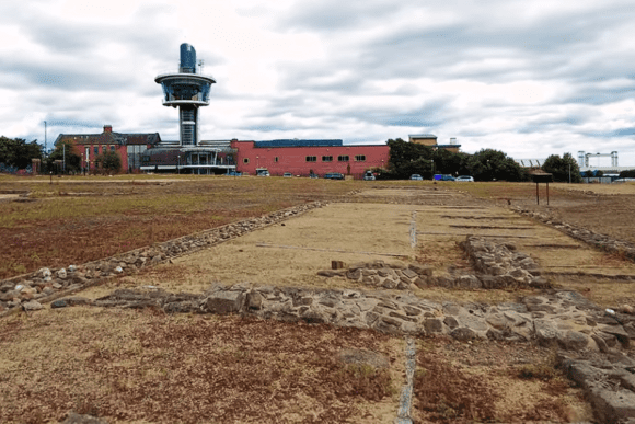 Ground-level view of Segedunum Roman Fort ruins with stone foundation outlines and modern visitor centre tower in the background, North Tyneside.
