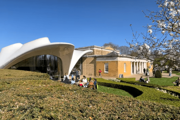Exterior view of Serpentine Gallery in Kensington Gardens, London, featuring the contemporary curved pavilion, landscaped gardens, blooming trees, and visitors enjoying the outdoor space on a sunny day.