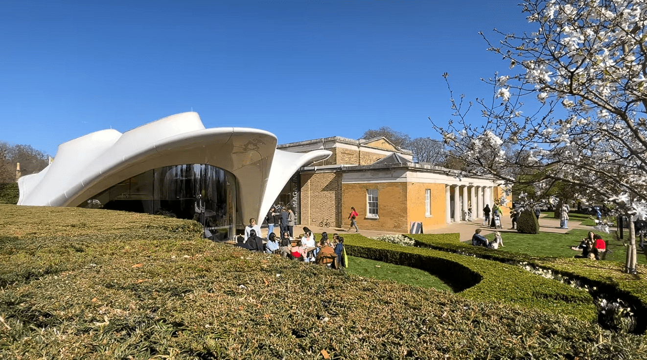 Exterior view of Serpentine Gallery in Kensington Gardens, London, featuring the contemporary curved pavilion, landscaped gardens, blooming trees, and visitors enjoying the outdoor space on a sunny day.