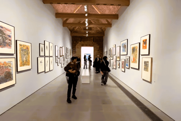 Visitors viewing framed contemporary artworks inside the Serpentine Galleries in London, displayed along a bright exhibition hall with high vaulted ceilings and minimalist gallery design.
