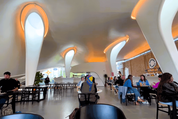 Interior of the Serpentine Pavilion café in Kensington Gardens, London, featuring futuristic curved white columns, warm ambient lighting, modern seating, and visitors dining within the iconic architectural space.