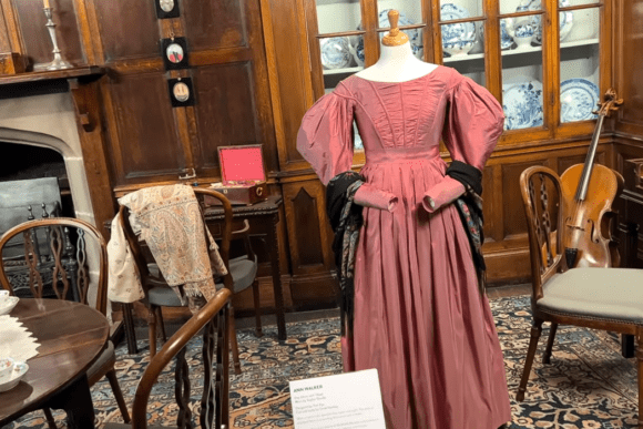 Victorian-era dress display at Shibden Hall with antique furniture, fireplace, and period interior setting in Halifax.