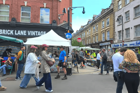 Busy Brick Lane street market in Shoreditch, London, with Brick Lane Vintage, market stalls, shoppers and historic East London buildings.
