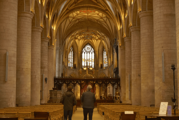 Interior nave of Tewkesbury Abbey featuring towering Norman columns, vaulted Gothic ceiling, stained glass windows, and visitors walking through the church.