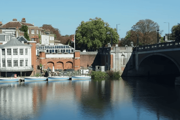 Scenic view along the Thames Path featuring riverside buildings, moored boats, a historic bridge, and calm reflections on the River Thames under a clear blue sky.