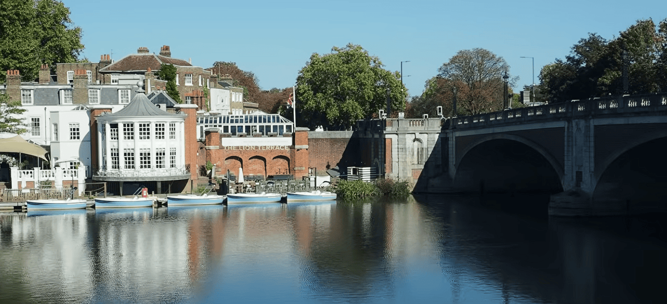 Scenic view along the Thames Path featuring riverside buildings, moored boats, a historic bridge, and calm reflections on the River Thames under a clear blue sky.