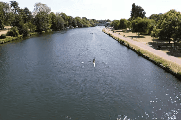 Aerial view of a lone rower on the River Thames beside the Thames Path, with tree-lined riverbanks, walking trails, and peaceful parkland on a sunny day.