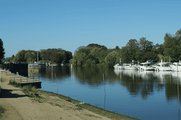 Peaceful stretch of the Thames Path with calm River Thames waters, moored boats, riverside trees, and a scenic walking trail under clear blue skies.