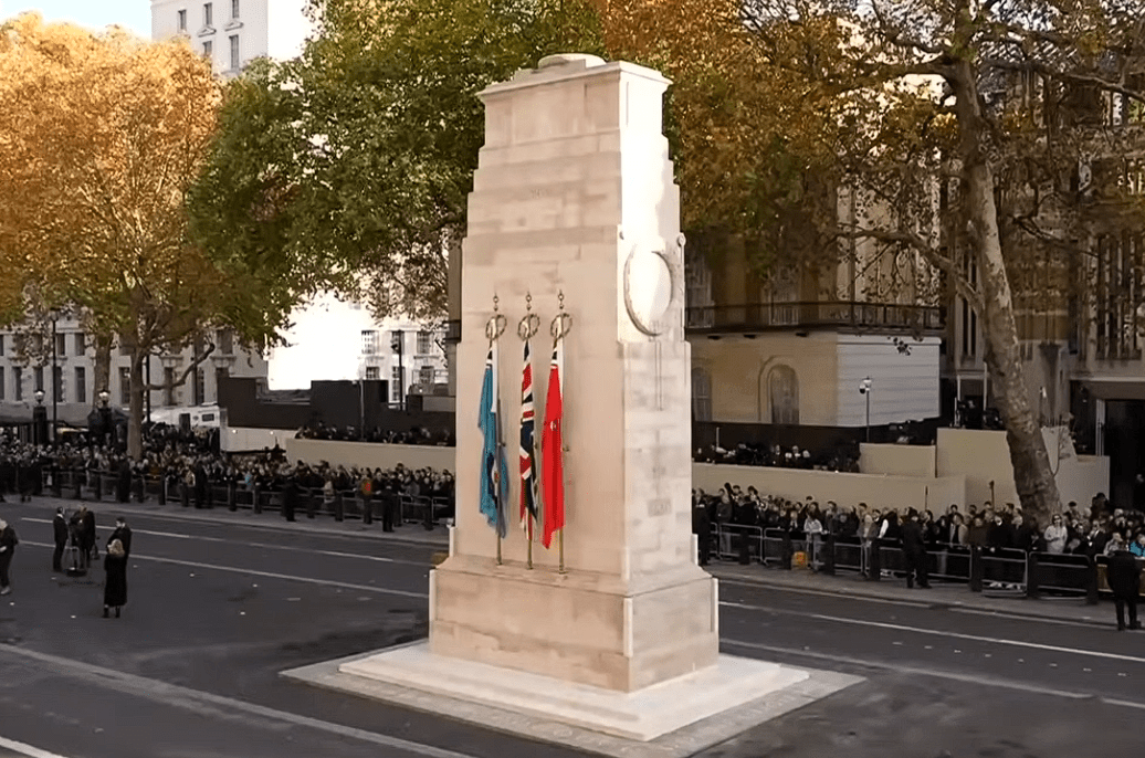 The Cenotaph war memorial in Whitehall, London, surrounded by crowds and ceremonial flags during a remembrance event.