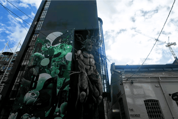 Large colourful street art murals covering a building at the Custard Factory in Digbeth, Birmingham, featuring green abstract designs and a dramatic human figure.