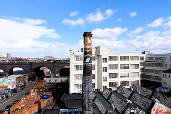 Aerial view of the Custard Factory in Digbeth, Birmingham, featuring historic industrial buildings, a tall brick chimney, railway arches and colourful street art.