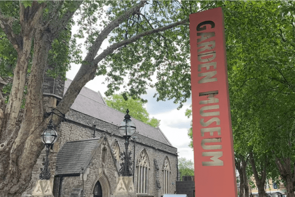 Entrance sign for The Garden Museum in London standing beside the historic church building, surrounded by mature trees and landscaped greenery near Lambeth Palace.