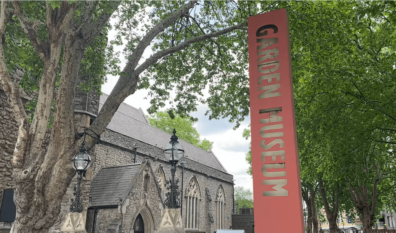 Entrance sign for The Garden Museum in London standing beside the historic church building, surrounded by mature trees and landscaped greenery near Lambeth Palace.