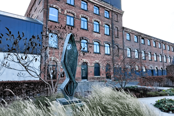 Outdoor sculpture set within landscaped gardens beside The Hepworth Wakefield, featuring historic red-brick mill architecture, contemporary public art, and the museum’s riverside cultural setting in West Yorkshire.