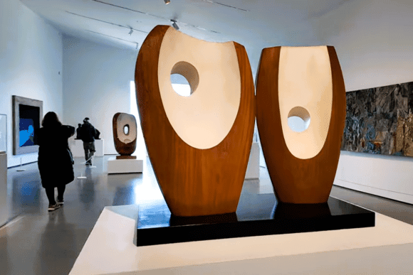 Abstract modern sculpture displayed inside The Hepworth Wakefield gallery, featuring large curved wooden forms with circular openings surrounded by contemporary paintings and artworks.