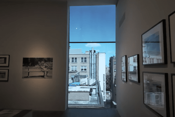 Photography exhibition gallery at The Photographers’ Gallery in London featuring framed photographic artworks displayed beside a large floor-to-ceiling window overlooking Soho, creating a blend of urban views and contemporary visual art.