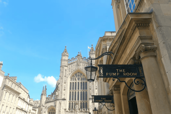 Exterior of The Pump Room Restaurant in Bath with its elegant Georgian entrance sign and the magnificent Bath Abbey rising in the background beneath a clear blue sky.