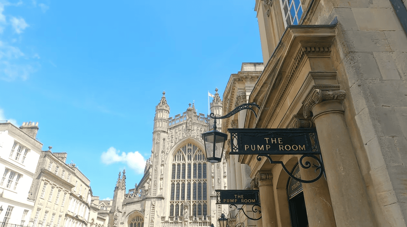 Exterior of The Pump Room Restaurant in Bath with its elegant Georgian entrance sign and the magnificent Bath Abbey rising in the background beneath a clear blue sky.