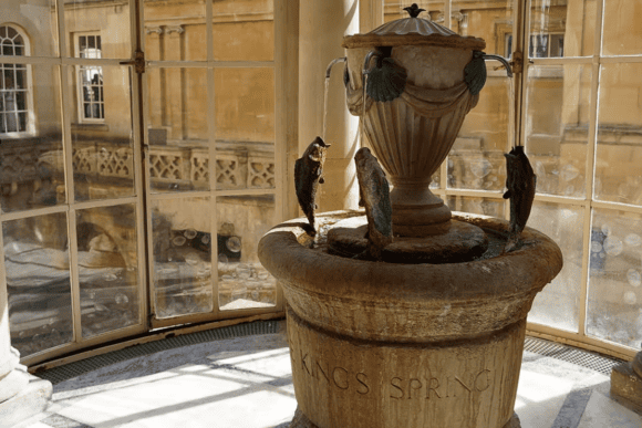 Historic King's Spring fountain inside The Pump Room Restaurant in Bath, featuring a decorative stone fountain with fish spouts overlooking the ancient Roman Baths through large Georgian windows.
