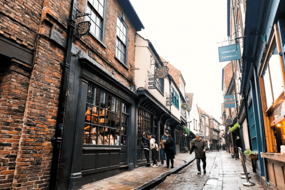 Visitors walking along The Shambles in York beside historic brick and timber-framed shops on the famous medieval cobbled street.