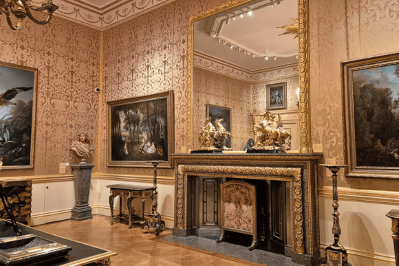 Elegant period room inside The Wallace Collection in London featuring ornate wallpaper, a gilded fireplace, antique furniture, sculptures, and framed Old Master paintings displayed in a historic gallery setting.