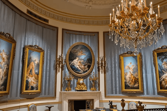 Elegant gallery room at The Wallace Collection in London featuring Rococo paintings in ornate gilded frames, a crystal chandelier, decorative mantelpiece, and luxurious historic interiors.