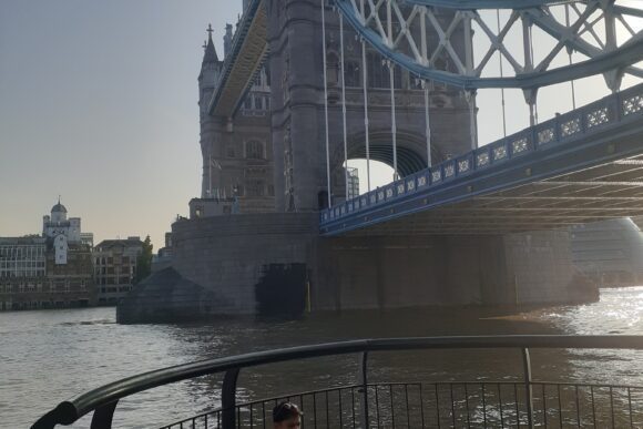 Tower Bridge in London viewed from the riverside walkway on the River Thames, with the iconic bridge tower and suspension structure in sunlight.