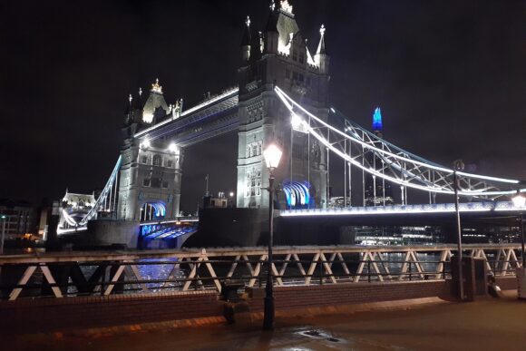 Tower Bridge illuminated at night over the River Thames in London, featuring glowing suspension cables and Gothic towers reflected on the water.