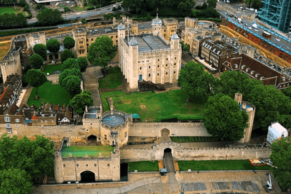 Aerial view of the Tower of London featuring the White Tower, medieval walls, fortified towers, and surrounding historic grounds beside the River Thames.