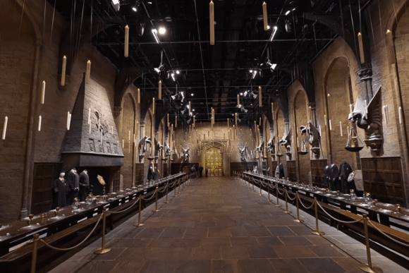 The Great Hall set at Warner Bros. Studio Tour London – The Making of Harry Potter, featuring long dining tables, floating candles, house costumes, and the iconic golden doors from Hogwarts School of Witchcraft and Wizardry.