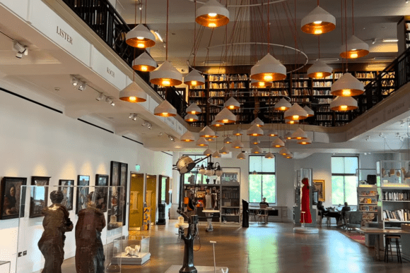 Interior of the Wellcome Collection in London featuring a spacious gallery and library area with hanging pendant lights, bookshelves, medical history exhibits, artworks, and visitors exploring the cultural space.