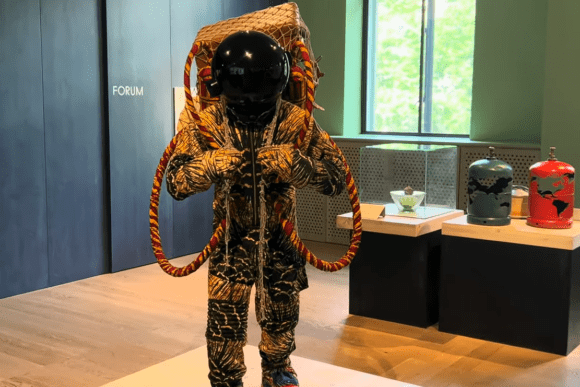 Contemporary astronaut-inspired sculpture on display at the Wellcome Collection in London, featuring a reflective black helmet, textured suit, and artistic life-support elements within a modern gallery exhibition.