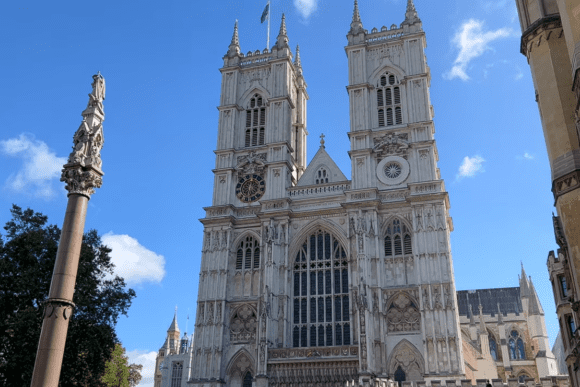 Front view of Westminster Abbey in London featuring its iconic Gothic twin towers, ornate stone façade, and historic architecture beneath a bright blue sky.