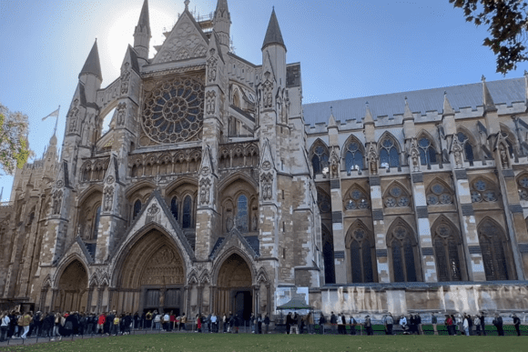 Side view of Westminster Abbey in London featuring Gothic arches, stained-glass rose window, ornate stone carvings, and visitors gathered outside the historic cathedral.