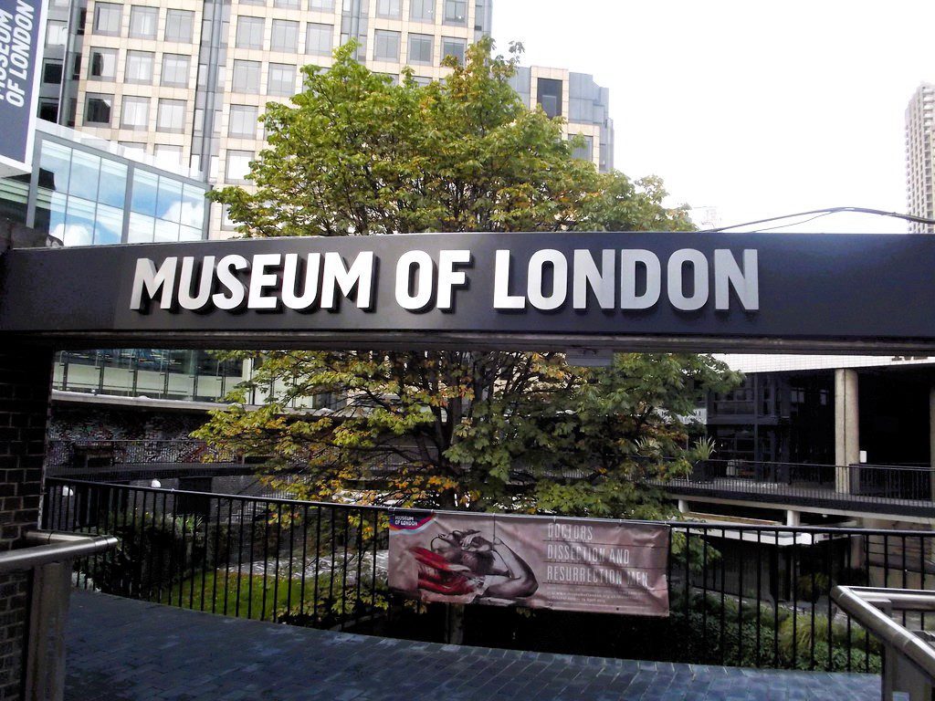 Museum of London entrance sign displayed outside the museum in central London, surrounded by modern city architecture and urban greenery.
