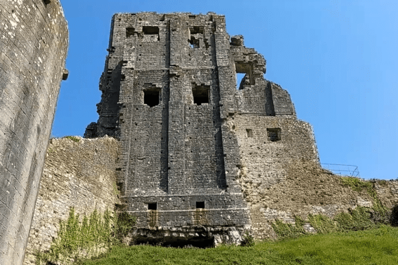 The ruined central keep of Corfe Castle rising above stone walls and grassy slopes under a clear blue sky.