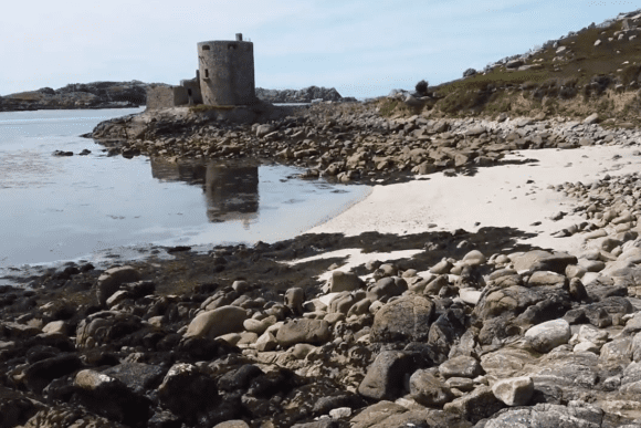 Cromwell’s Castle reflected in calm coastal water beside a rocky beach on Tresco.