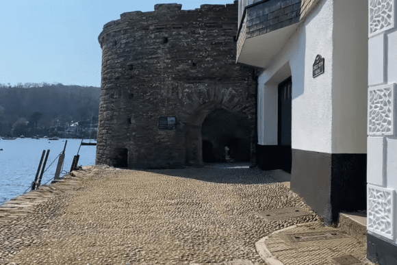 Stone tower and arched entrance of Dartmouth Castle beside the River Dart with waterfront path in the foreground.