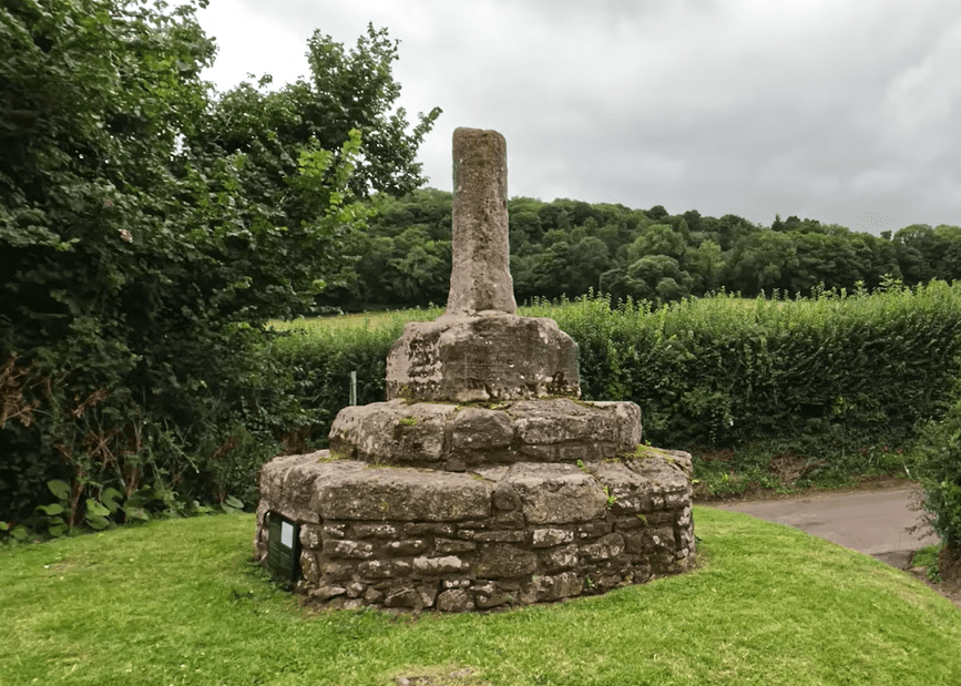Stone Butter Cross monument at Dunster, standing on a stepped circular base in a grassy village setting.