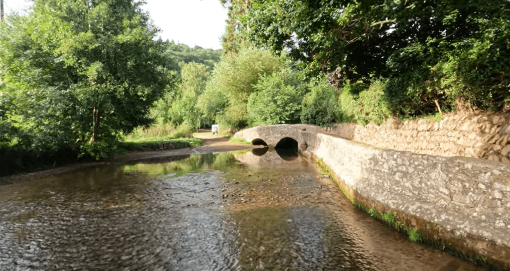 Stone packhorse bridge with twin arches crossing a shallow river at Dunster Gallox Bridge, surrounded by trees.