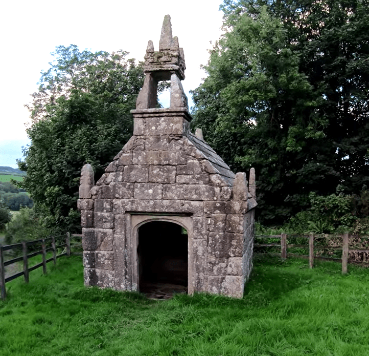 Dupath Well, a small stone well house with a stepped roof and carved finial, set in a grassy clearing.