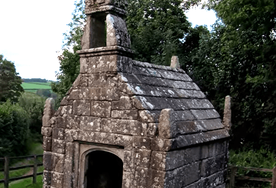 Stone well house at Dupath Well with a stepped slate roof, arched doorway, and tall carved finial, surrounded by grass and trees.