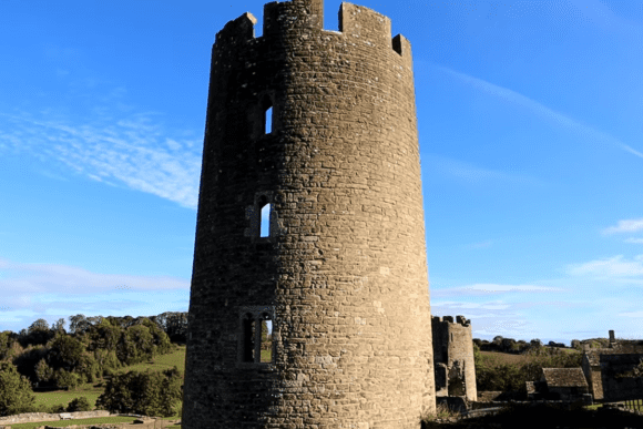 Round stone tower with narrow slit windows at Farleigh Hungerford Castle, rising above the countryside under a clear blue sky.