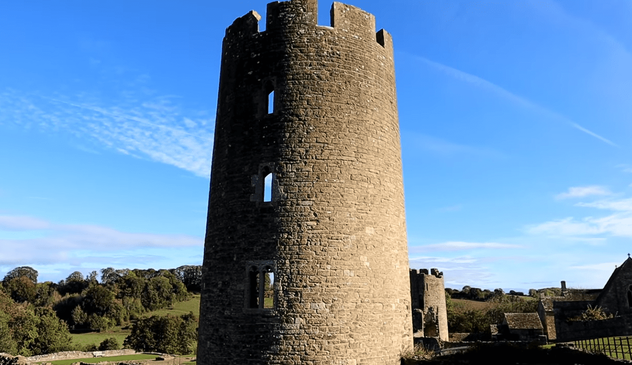 Round stone tower with narrow slit windows at Farleigh Hungerford Castle, rising above the countryside under a clear blue sky.