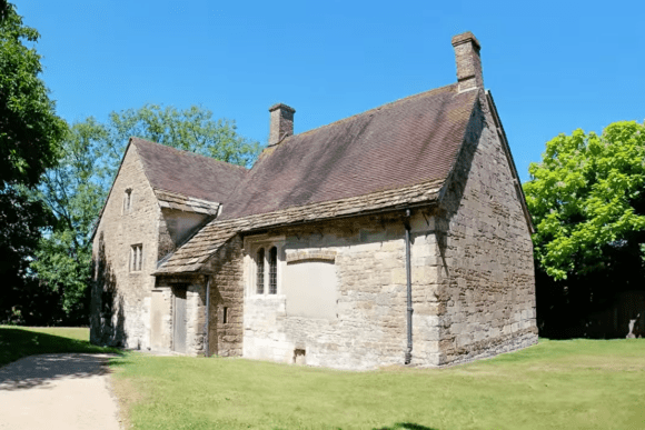 Stone-built medieval house at Fiddleford Manor with a steep tiled roof, chimneys, and small leaded windows, set among trees on a sunny day.