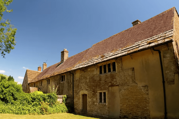 Long stone range of Fiddleford Manor with a steep tiled roof, small mullioned windows, and chimneys, set in a grassy garden under a clear blue sky.