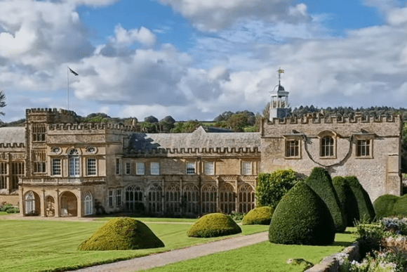 Wide view of Forde Abbey with its historic stone buildings, formal lawns, and manicured gardens under a partly cloudy sky.