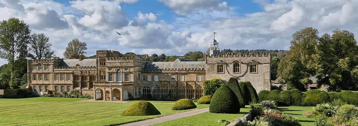 Wide view of Forde Abbey with its historic stone buildings, formal lawns, and manicured gardens under a partly cloudy sky.