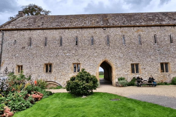 Stone façade of Forde Abbey with an arched gateway, small windows, flower beds, and visitors seated on benches.